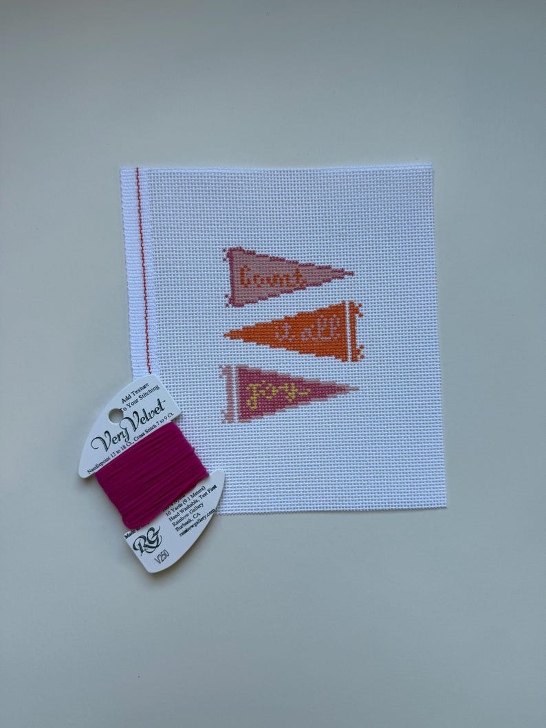 K&ouml;nnte beinhalten: Eine Gobelin-Leinwand mit drei Wimpeln, die in Orange- und Rosat&ouml;nen gestickt sind, jeweils mit einem Wort: "Count", "it all" und "joy". Eine Spule rosa Very Velvet-Garn befindet sich in der linken unteren Ecke.