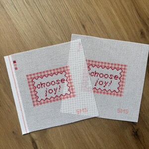May include: Two square pieces of white needlepoint canvas featuring a red gingham border and the text "choose joy!" in red cross-stitch lettering. Each canvas has a small red vertical line on the left edge and the letters "SMS" in red.