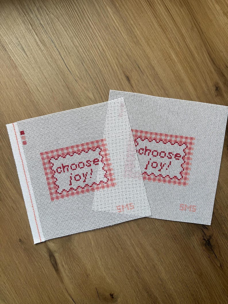 May include: Two square pieces of white needlepoint canvas featuring a red gingham border and the text "choose joy!" in red cross-stitch lettering. Each canvas has a small red vertical line on the left edge and the letters "SMS" in red.