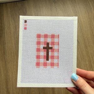 May include: A needlepoint canvas with a brown cross on a pink and white checked pattern. The canvas has a white border and is held by a hand with blue nail polish.