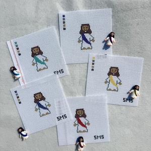 May include: Five square white needlepoint canvases featuring a pixelated figure of a person with brown hair and a robe. Each canvas displays a different colored sash: teal, blue, yellow, red, and purple. Each canvas includes the text SMS.