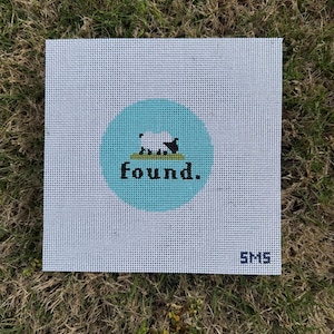 May include: A square white needlepoint canvas featuring a central light blue circle. Inside the circle is a small black and white sheep standing on a green patch of grass above the black text "found." The letters "SMS" are printed in the bottom right corner.