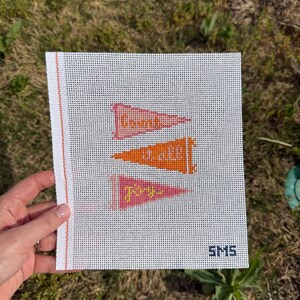 K&ouml;nnte beinhalten: Eine wei&szlig;e Leinwand mit drei Wimpelfahnen in Rosa, Orange und Rosa, jeweils mit Text. Die Fahnen sind mit Kreuzstich versehen. Die Leinwand wird von einer Hand gehalten und tr&auml;gt die Initialen "SMS" unten rechts.