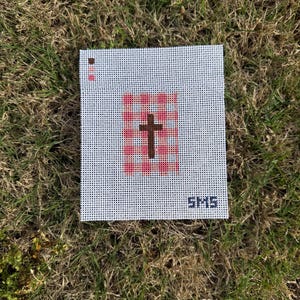 May include: A square needlepoint canvas with a white background and a grid pattern. The design features a brown cross in the center, surrounded by pink shapes. The letters "SMS" are in the bottom right corner.