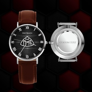 May include: A Maybach wristwatch with a black face, silver accents, and brown leather band. The watch face features Roman numerals and the Maybach logo. The back of the watch is silver with the words "CUSTOM TEXT".