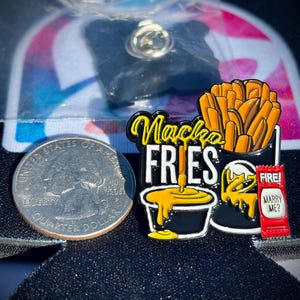 May include: A metal enamel pin featuring a container of orange fries, a black cup of yellow cheese sauce, and a red sauce packet labeled FIRE! and MARRY ME? next to a US quarter for scale. The text Nacho FRIES is written in yellow and white.