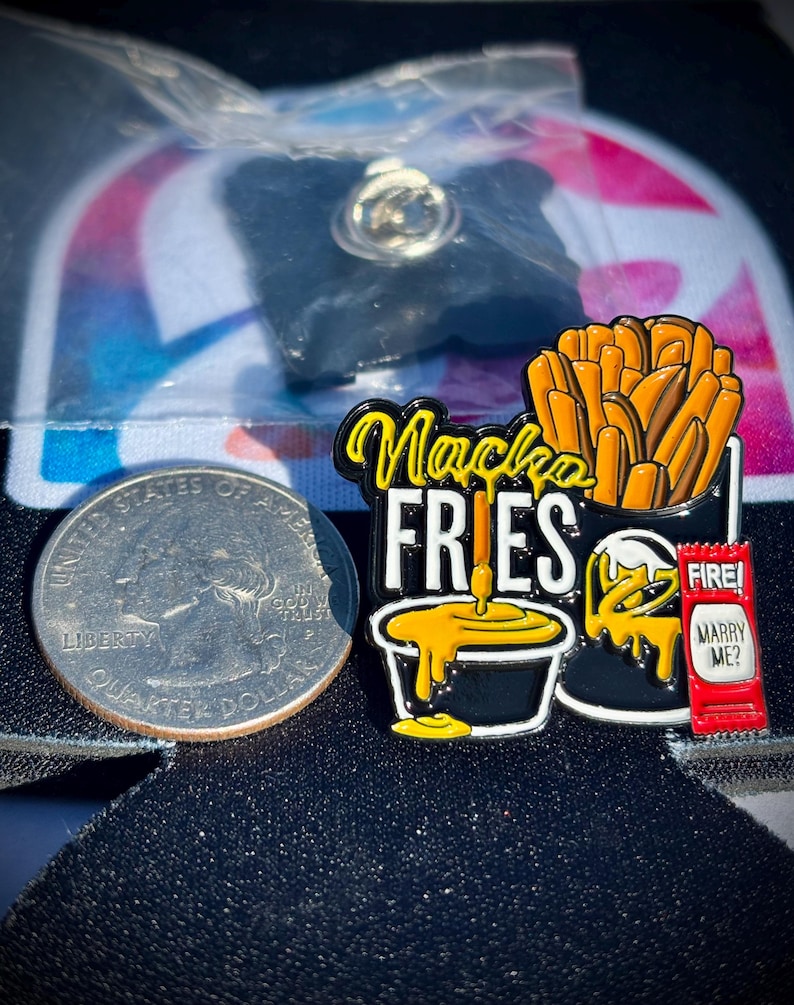 May include: A metal enamel pin featuring a container of orange fries, a black cup of yellow cheese sauce, and a red sauce packet labeled FIRE! and MARRY ME? next to a US quarter for scale. The text Nacho FRIES is written in yellow and white.