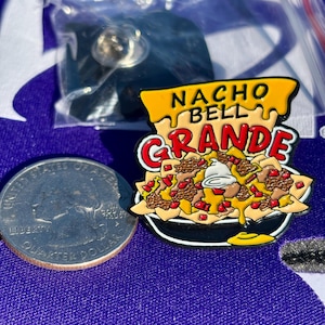May include: A metal enamel pin shaped like a bowl of nachos with the text NACHO BELL GRANDE in yellow and red. The pin features yellow chips, brown ground meat, red diced tomatoes, and white sour cream. A US quarter is shown for scale.