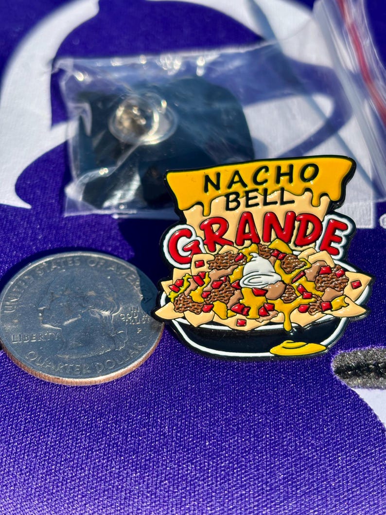 May include: A metal enamel pin shaped like a bowl of nachos with the text NACHO BELL GRANDE in yellow and red. The pin features yellow chips, brown ground meat, red diced tomatoes, and white sour cream. A US quarter is shown for scale.