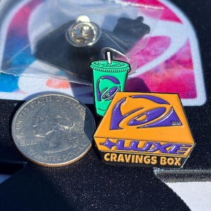 May include: Two enamel pins featuring a green drink cup with a purple logo and an orange trapezoidal box with a purple logo and the text LUXE CRAVINGS BOX, displayed next to a United States quarter for scale, with one pin inside a plastic bag.