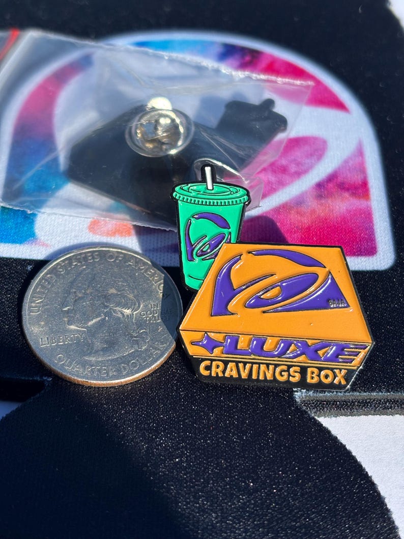May include: Two enamel pins featuring a green drink cup with a purple logo and an orange trapezoidal box with a purple logo and the text LUXE CRAVINGS BOX, displayed next to a United States quarter for scale, with one pin inside a plastic bag.