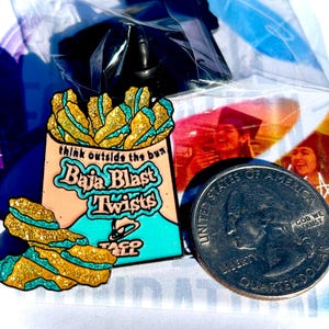 May include: A metal enamel pin shaped like a bag of Baja Blast Twists with gold glitter and teal accents. The bag features the text 'think outside the bun', 'Baja Blast Twists', and the Taco Bell logo. A US quarter is placed next to the pin for scale.