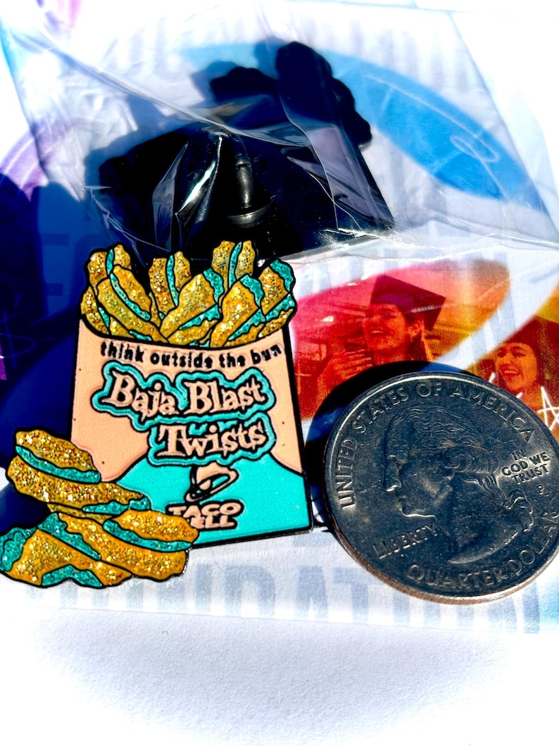 May include: A metal enamel pin shaped like a bag of Baja Blast Twists with gold glitter and teal accents. The bag features the text 'think outside the bun', 'Baja Blast Twists', and the Taco Bell logo. A US quarter is placed next to the pin for scale.