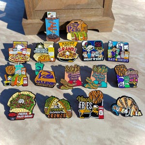 May include: A collection of colorful enamel pins featuring various fast food items, including tacos, burritos, fries, and branded meal boxes. Each pin displays detailed illustrations of food with text labels like Mexican Pizza, Nacho Bell Grande, and Quesadilla.