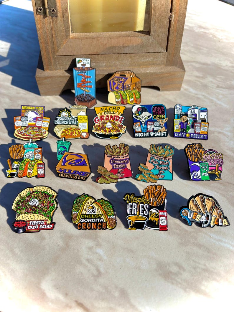 May include: A collection of colorful enamel pins featuring various fast food items, including tacos, burritos, fries, and branded meal boxes. Each pin displays detailed illustrations of food with text labels like Mexican Pizza, Nacho Bell Grande, and Quesadilla.