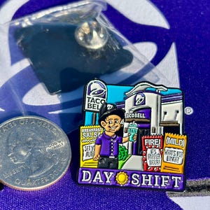 May include: A Taco Bell enamel pin featuring a worker in a purple shirt holding a drink in front of a restaurant. The pin includes text reading Breakfast Salsa, Fire, Mild, and Day Shift with a sun icon. A US quarter is shown for scale.