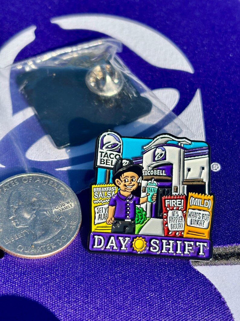 May include: A Taco Bell enamel pin featuring a worker in a purple shirt holding a drink in front of a restaurant. The pin includes text reading Breakfast Salsa, Fire, Mild, and Day Shift with a sun icon. A US quarter is shown for scale.