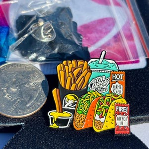 May include: A metal enamel pin featuring a fast food meal including a container of golden fries, a teal drink cup labeled Mtn Dew Baja Blast, two tacos, a small black cup with yellow dipping sauce, and two red sauce packets labeled HOT GAME OVER and FIRE! GO FOR IT.