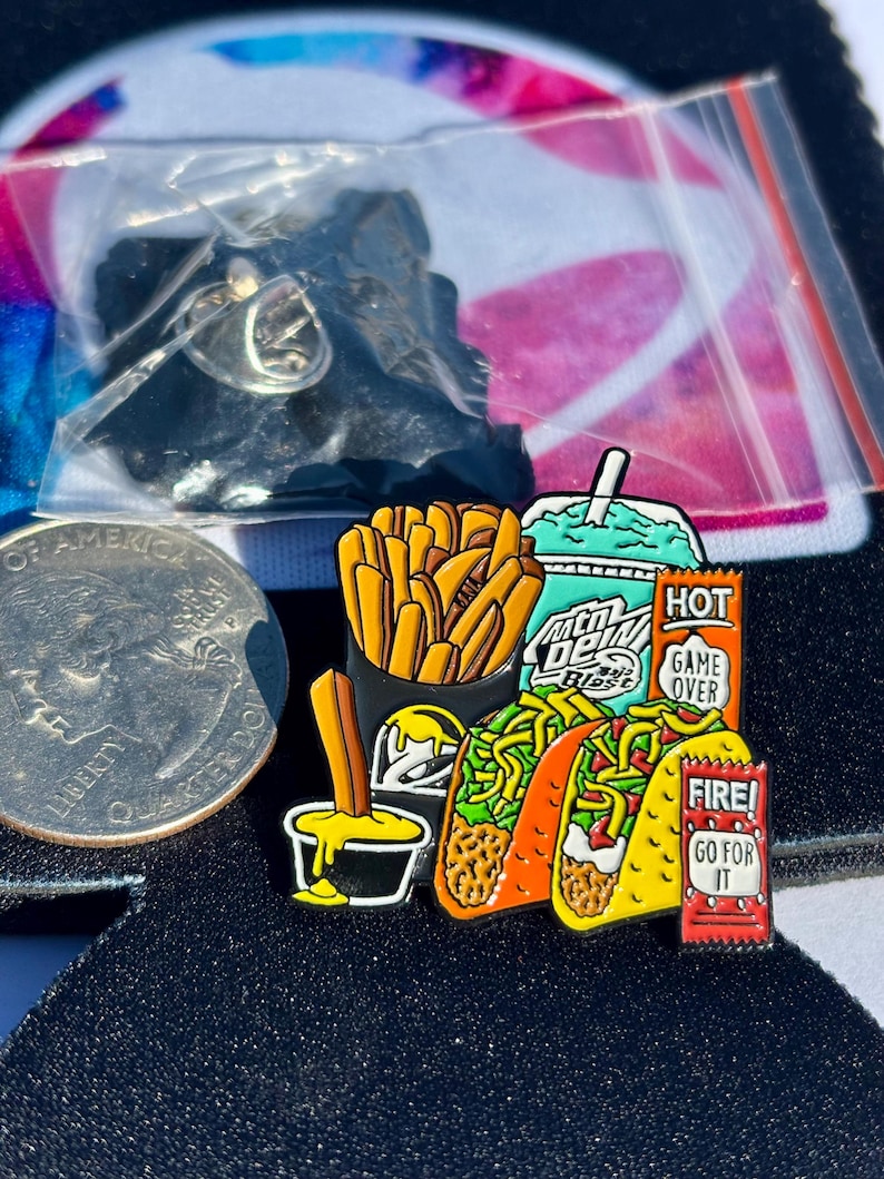 May include: A metal enamel pin featuring a fast food meal including a container of golden fries, a teal drink cup labeled Mtn Dew Baja Blast, two tacos, a small black cup with yellow dipping sauce, and two red sauce packets labeled HOT GAME OVER and FIRE! GO FOR IT.