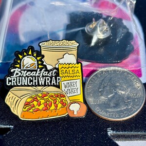 May include: A metal enamel pin featuring a breakfast crunchwrap, a cup, and a salsa packet with the text Breakfast CRUNCHWRAP and WAKEY WAKEY. The pin is positioned next to a United States quarter dollar coin for scale, showing the size of the item.