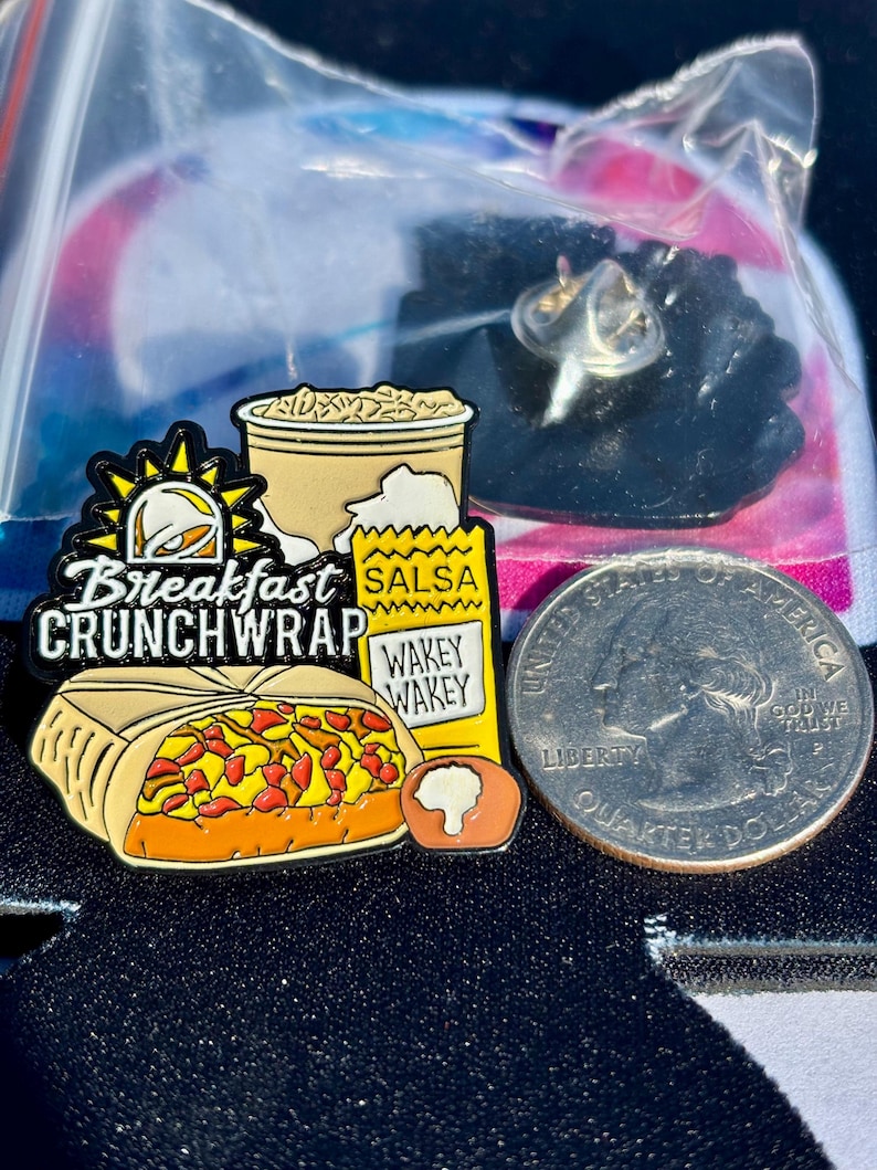 May include: A metal enamel pin featuring a breakfast crunchwrap, a cup, and a salsa packet with the text Breakfast CRUNCHWRAP and WAKEY WAKEY. The pin is positioned next to a United States quarter dollar coin for scale, showing the size of the item.