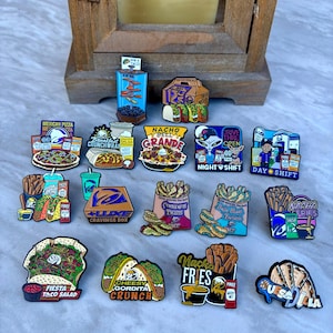 May include: A collection of colorful enamel pins featuring various Taco Bell food items, including a Mexican Pizza, Crunchwrap, Nacho BellGrande, and various fries, with text labels like 'Nacho Fries', 'Fiesta Taco Salad', and 'Cheesy Gordita Crunch'.