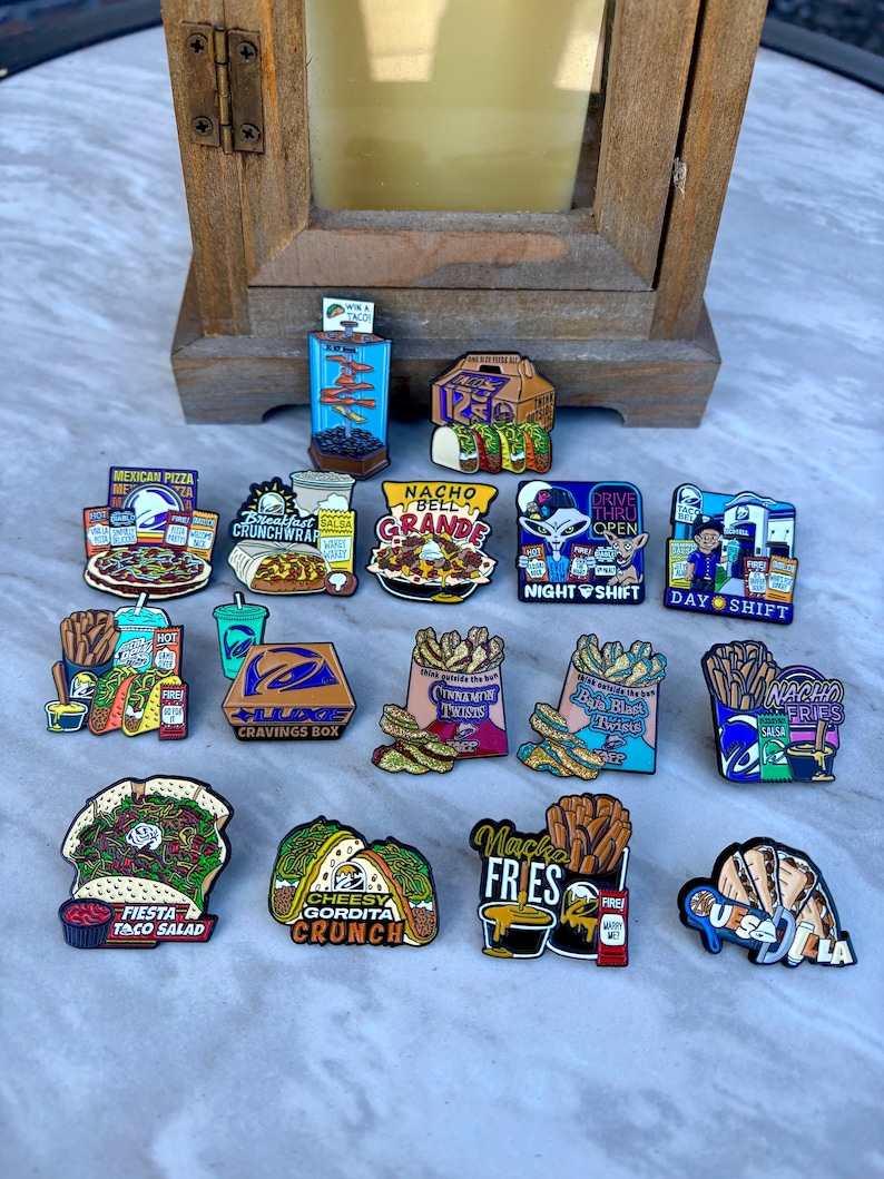 May include: A collection of colorful enamel pins featuring various Taco Bell food items, including a Mexican Pizza, Crunchwrap, Nacho BellGrande, and various fries, with text labels like 'Nacho Fries', 'Fiesta Taco Salad', and 'Cheesy Gordita Crunch'.