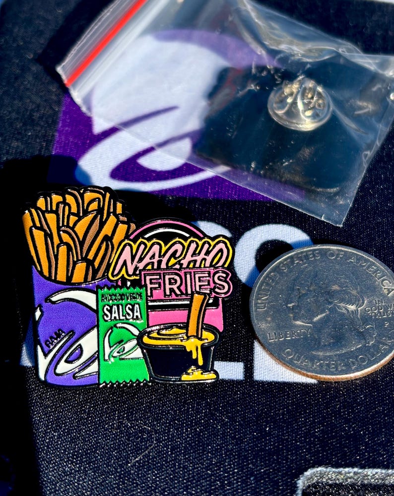 May include: A metal enamel pin featuring a purple container of golden brown fries, a green packet labeled Salsa, and a black cup of yellow cheese sauce with a fry dipped in it. The text Nacho Fries is in pink. A US quarter is shown for scale.