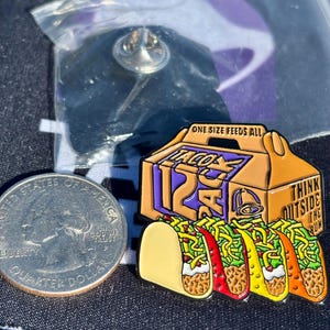 May include: A metal enamel pin featuring a Taco Bell 12-pack box with the text ONE SIZE FEEDS ALL and THINK OUTSIDE THE BUN, positioned next to a United States quarter dollar coin for scale. The pin depicts four tacos with lettuce, cheese, and meat.