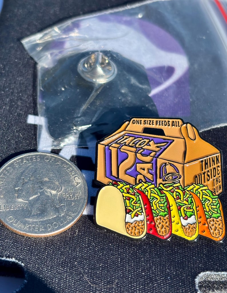 May include: A metal enamel pin featuring a Taco Bell 12-pack box with the text ONE SIZE FEEDS ALL and THINK OUTSIDE THE BUN, positioned next to a United States quarter dollar coin for scale. The pin depicts four tacos with lettuce, cheese, and meat.