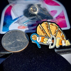 May include: A metal enamel pin featuring three folded quesadillas above the text QUESADILLA, with a blue tennis racket and a white bell integrated into the lettering, positioned next to a United States quarter for scale.