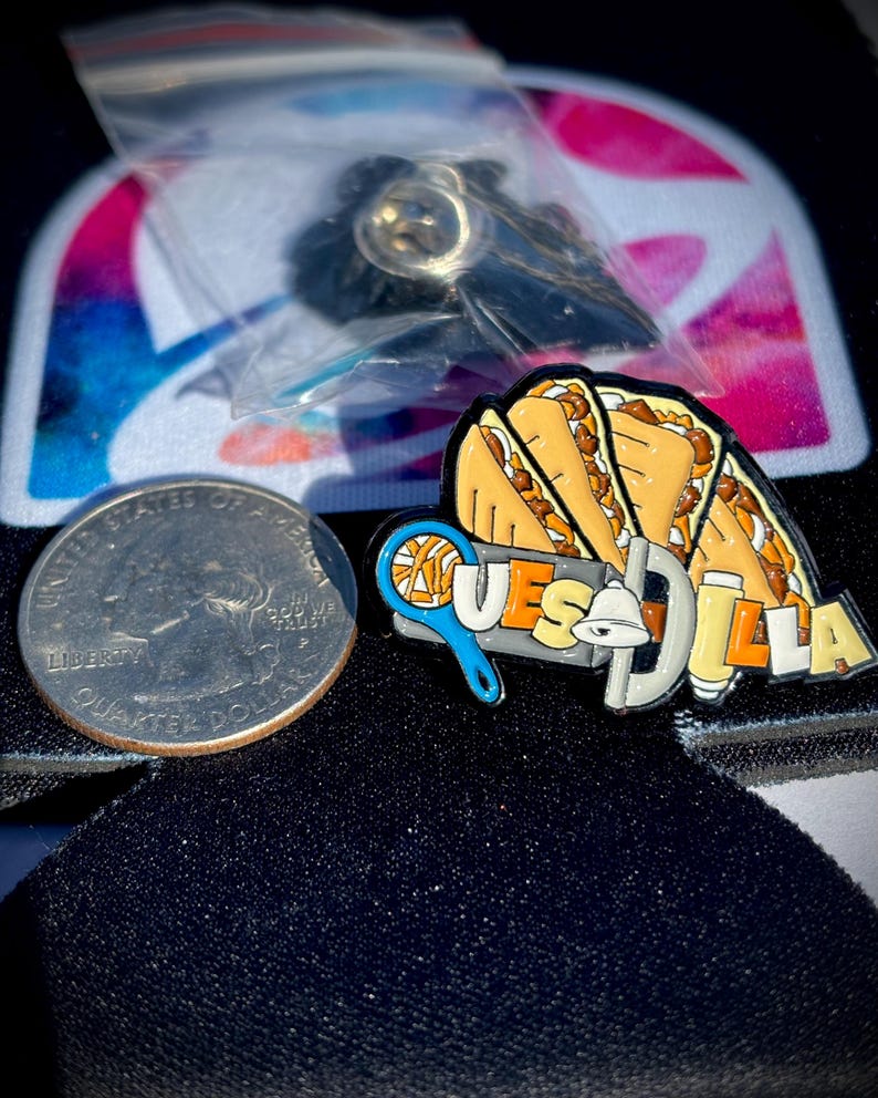 May include: A metal enamel pin featuring three folded quesadillas above the text QUESADILLA, with a blue tennis racket and a white bell integrated into the lettering, positioned next to a United States quarter for scale.