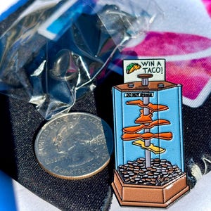May include: A metal enamel pin shaped like a coin pusher arcade machine with a brown base, blue sides, and a central pole holding orange shapes. The top sign reads WIN A TACO! and DO NOT SHAKE. A US quarter is placed next to the pin for scale reference.