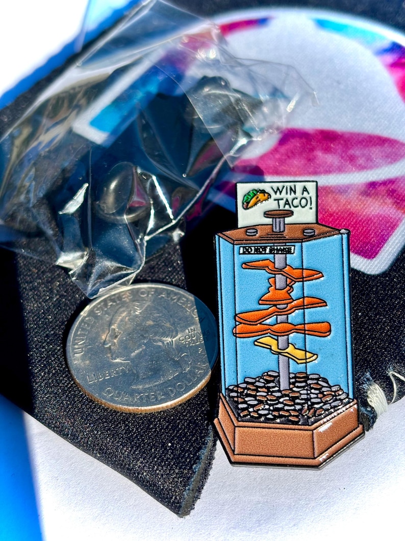 May include: A metal enamel pin shaped like a coin pusher arcade machine with a brown base, blue sides, and a central pole holding orange shapes. The top sign reads WIN A TACO! and DO NOT SHAKE. A US quarter is placed next to the pin for scale reference.