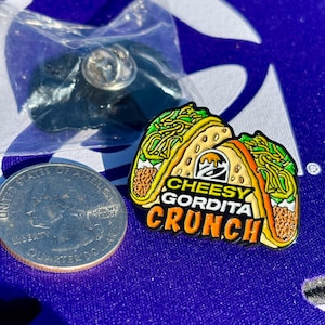 May include: A metal enamel pin shaped like a Cheesy Gordita Crunch taco with yellow, green, and brown details. The text reads CHEESY GORDITA CRUNCH with the Taco Bell logo. A US quarter coin is placed next to the pin for scale, and a plastic bag is visible.