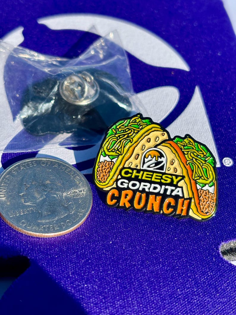 May include: A metal enamel pin shaped like a Cheesy Gordita Crunch taco with yellow, green, and brown details. The text reads CHEESY GORDITA CRUNCH with the Taco Bell logo. A US quarter coin is placed next to the pin for scale, and a plastic bag is visible.