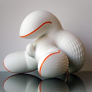 May include: A white abstract sculpture with orange detailing. The sculpture has a rounded form with textured details and orange lines outlining the edges. The artwork is displayed on a reflective surface.