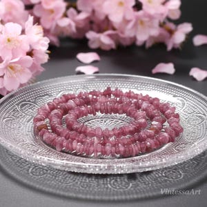 Könnte beinhalten: Eine Halskette aus kleinen, unregelmäßigen, rosavioletten Edelsteinen, spiralförmig auf einem durchsichtigen, gemusterten Glasteller angeordnet. Rosa Blüten und Blütenblätter sind im Hintergrund. Der Text "VintessaArt" befindet sich unten rechts.