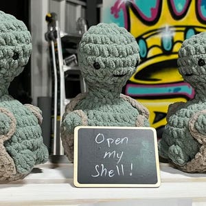 May include: Three handmade crochet turtle plushies in shades of green and tan. Each turtle has a round head, small black eyes, and a shell-like body. A small chalkboard in front of the turtles reads "Open my Shell!"