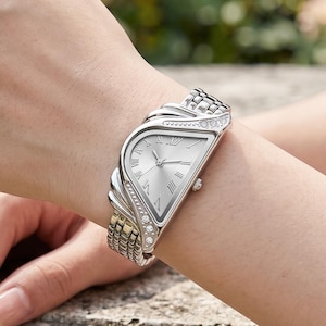Op de afbeelding: Een zilverkleurig polshorloge met een uniek, asymmetrisch ontwerp. Het horloge heeft een zilveren wijzerplaat met Romeinse cijfers en een decoratieve band met heldere kristallen. De band is een mix van zilver- en goudkleurig metaal.