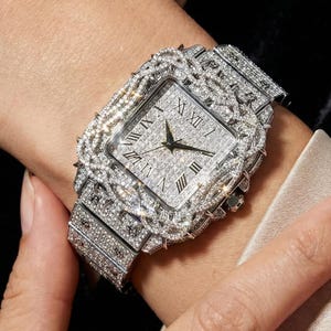 Op de afbeelding: Een luxe horloge met een rechthoekige wijzerplaat en een met diamanten bezette band. De wijzerplaat heeft Romeinse cijfers. Het horloge wordt om de pols gedragen, wat het ingewikkelde ontwerp en de luxueuze uitstraling benadrukt.
