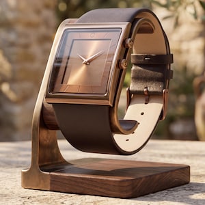 Puede incluir: Un reloj de esfera cuadrada con correa de cuero marrón y esfera color cobre. El reloj se exhibe sobre un soporte de madera con marco de metal color cobre. La esfera del reloj tiene un diseño minimalista con manecillas finas.