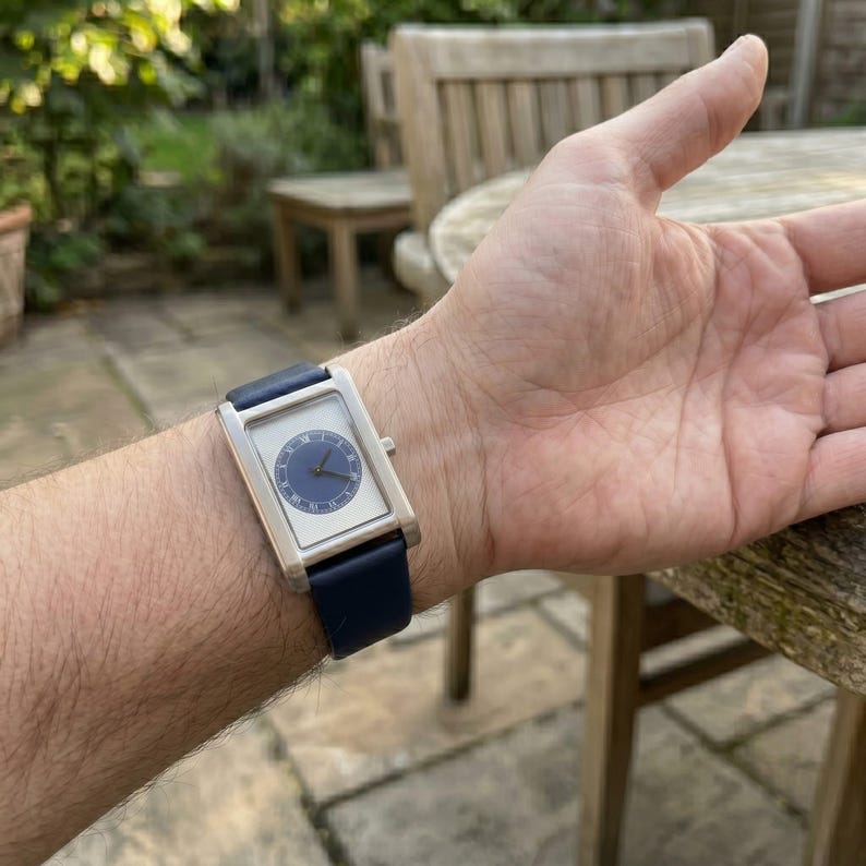 May include: A rectangular wristwatch with a silver case and a navy blue leather band. The watch face is white with a blue circular dial and black hands. The watch is on a wrist.