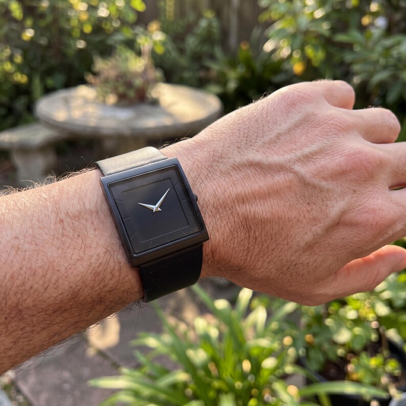 May include: A black square wristwatch with a black leather strap and a light grey strap. The watch face is black with silver hands. The watch is on a wrist.