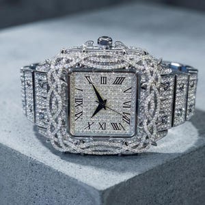 Op de afbeelding: Een luxe horloge met een vierkante wijzerplaat en een zilveren band. Het horloge is bezet met talrijke kleine, sprankelende diamanten. Romeinse cijfers markeren de uren op de wijzerplaat. Het horloge ligt op een grijze ondergrond.