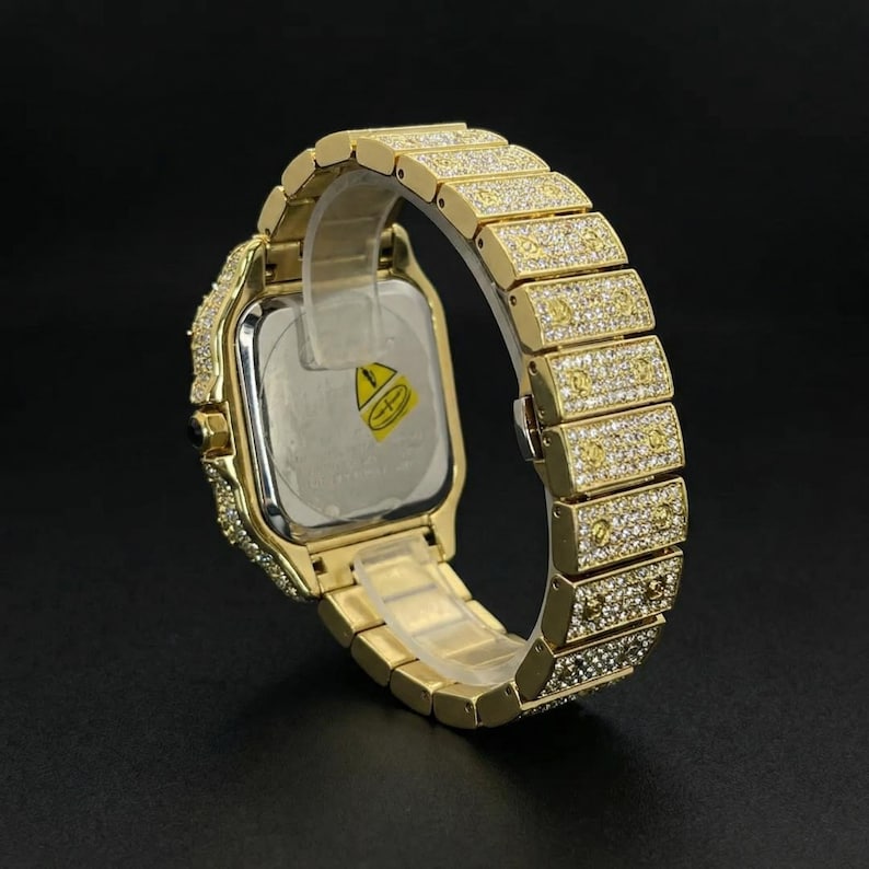 Op de afbeelding: Goudkleurig polshorloge met een vierkante wijzerplaat en een band bezet met sprankelende stenen. Het horloge heeft een goudkleurige metalen band en een doorzichtig plastic stuk dat het horloge op zijn plaats houdt. De wijzerplaat is zilverkleurig met een gele en groene sticker.