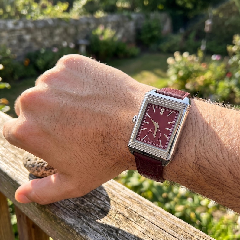 May include: A rectangular wristwatch with a burgundy face and a silver-colored case. The watch has a burgundy leather band. The watch is on a wrist, with a wooden fence and greenery in the background.