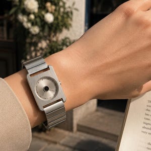 Puede incluir: Un reloj de pulsera plateado con una esfera rectangular y una correa de metal a juego. La esfera del reloj tiene un diseño circular con un centro negro. El reloj se lleva en la muñeca.