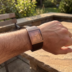 May include: A rose gold square watch with a dark brown leather strap. The watch face is a light copper colour with gold hands. The watch is on a wrist, with the background out of focus.