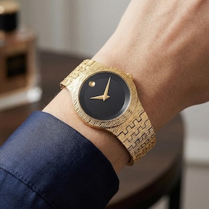 Puede incluir: Reloj de pulsera dorado con una correa texturizada y una esfera negra. El reloj presenta agujas doradas y un pequeño punto a las 12 en punto. El reloj está en una muñeca, con una manga azul marino visible.